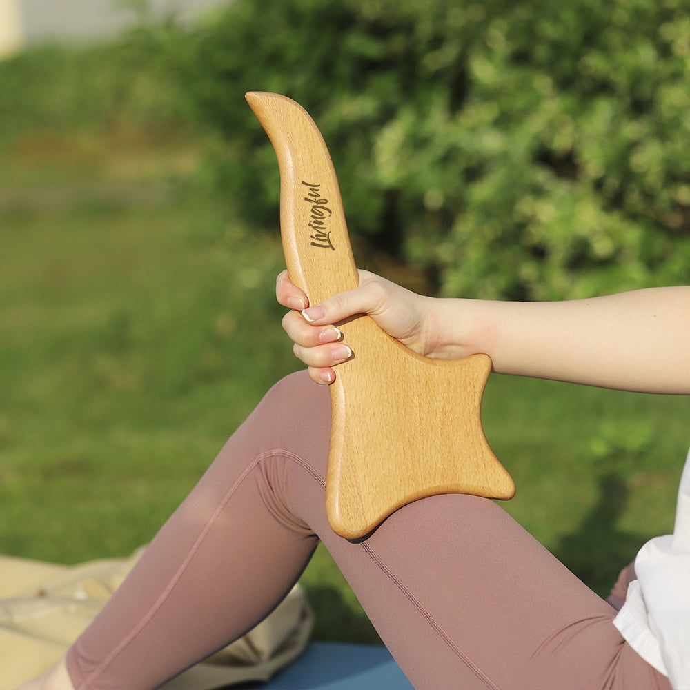 A person holding the Premium Wood Massage Tool 4 Sides by Livingful, featuring an elongated handle and a flat, star-shaped base, sitting outdoors surrounded by greenery.