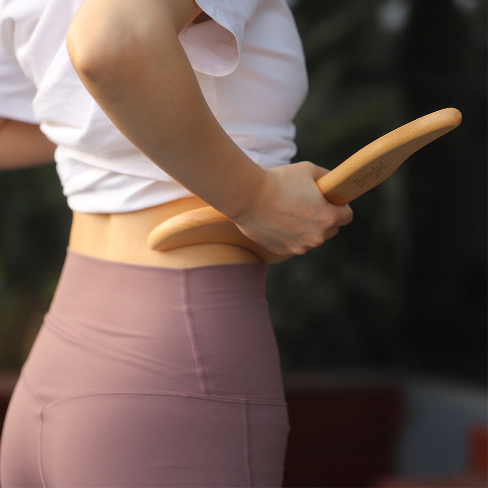 A person in a white shirt and pink leggings uses the Livingful Premium Wood Massage Tool 3 Sides on their lower back, enjoying the soothing effects of a Gua Sha massage.