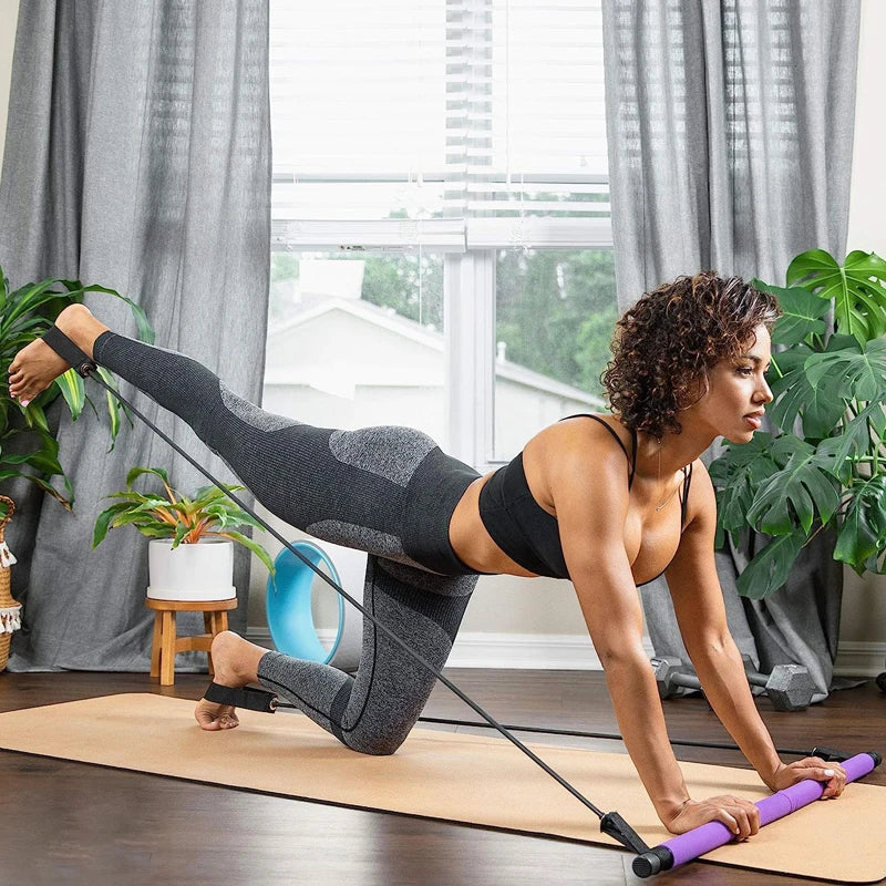 An individual is working out indoors with a resistance band, kneeling on a yoga mat and stretching one leg backward. The bright room, adorned with flourishing plants by the window, provides a tranquil setting ideal for using the IPCBEC Multifunctional Pilates Bar to boost strength and flexibility.